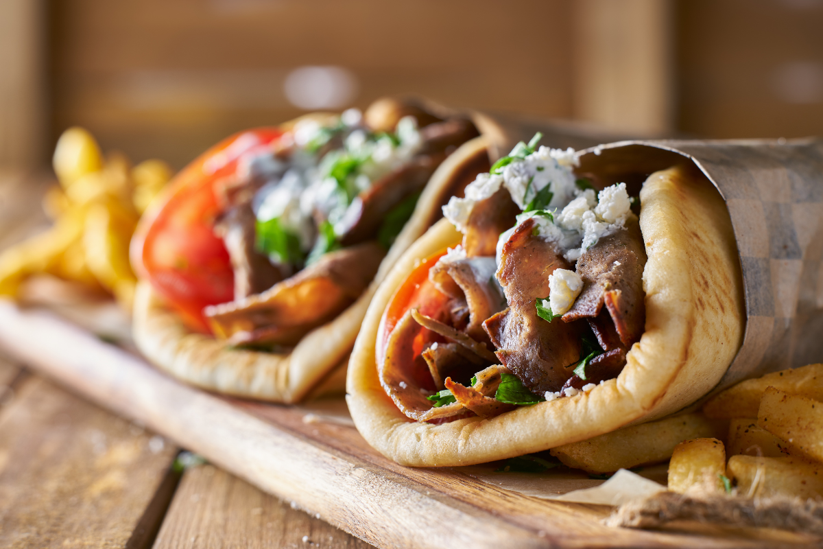 Two Greek pita gyros filled with sliced meat, tomatoes, herbs, and crumbled cheese, served with a side of fries on a wooden board.